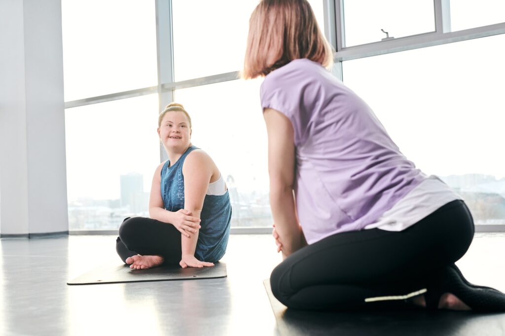 Happy disable girl in activewear looking at friend while both sitting on mats